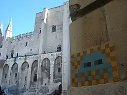 Photograph of the sides of two stone buildings, with a tan building close up in the foreground and a white one in the background. On the tan building is a mosaic made of colored tiles that form a pixelated "Space Invaders" alien.