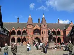 Stables, Eaton Hall, is directly north of the Chapel, in red brick, showing French late Gothic influence and the use of Tudor style half-timbering in the upper storey in the flanking ranges