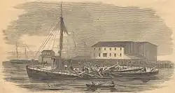 a black and white image of a wooden steamboat lying low in the water near a shore with buildings. The smokestack of the steam engine lies flat in a mass of broken planks in the center of the boat while men clamber about the wreckage. Three men in a rowboat observe the scene.