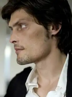 Unshaven, dark-haired young man looking left