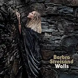 Barbra Streisand wears a black shall, gazing upward, surrounded by a large stone wall
