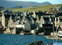 Stone houses crowd around a shore, the gable ends facing the water, with green hills beyond.