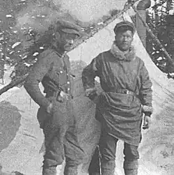In a grayscale photograph, two men stand in front of a tent and snowy evergreen trees
