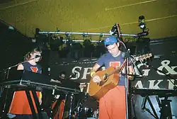 A man plays acoustic guitar in the centre of a stage, the black back wall of which is filled with the words "Spring & Airbrake" in white lettering. A female keyboardist is behind and to the left of the man, and in the background can be seen a male bassist and a drum kit.