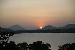A partial view of a large lake, seen over a tree line. The further bank is covered by a thick forest, and the sun is setting over a mountain range in the background.