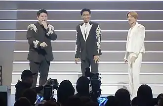 Three man standing on stage, wearing suits and wireless microphone headsets