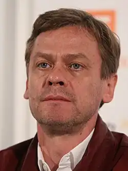 close-up of Sylvester Groth wearing a white shirt with a dark red jacket, looking just right of camera