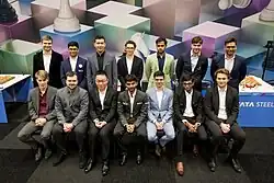 7 GMs sitting on chairs in a row, 7 standing behind them