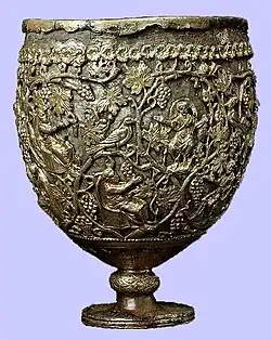 A photo of a large ovoid vessel standing on a short knobbed stem. The cup comprises a silver body enclosed in an openwork layer of gold. The gold ornamentation represents vine scrolls enclosing small seated and praying figures.