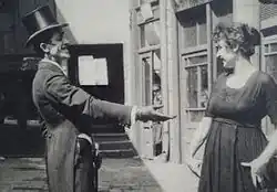 A man with a top hat and curled mustache reaching out his hand to a woman, both smiling, from a scene of The Bulgarian Is Gallant