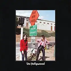 A photograph of two men standing by street signs is surrounded by a black border. In the center bottom, it reads: "Do Hollywood"