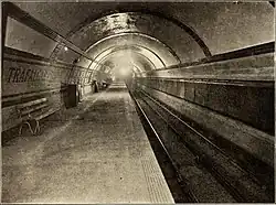 A circular tunnel with platform on the left and tracks on the right. The platform wall is tiled in bands with the words TRAFALGAR SQUARE picked out in coloured tiles
