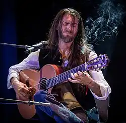 Estas Tonne sitting onstage, playing an acoustic guitar in front of a microphone, with a smoking incense stick attached to the guitar's neck