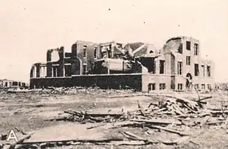 A school building, seen in black-and-white, is heavily damaged. At least three f its walls are crumbled, and the roof is missing.