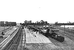 A black and white photo of a railway station from higher ground above the tracks a short distance away. There are four tracks with a platform either side; the right-hand platform is an island platform with a goods yard to the right. A train sits on the rightmost platform, which has many people on it.
