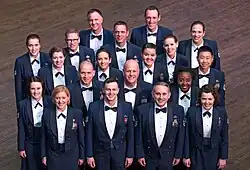 This is a picture of the 19 members of the official chorus of the United States Air Force, the Singing Sergeants. They are dressed in the dark blue Air Force mess dress uniform and are standing in 5 rows on a dark brown wood floor looking and smiling up at the photographer.