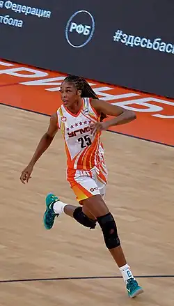Colour photograph of Victoria Macaulay age 33 playing basketball in a white and orange kit, knee braces, and blue Nike trainer/sneaker shoes.