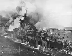Black and white photograph of a World War II-era aircraft carrier on fire. The portion of the ship rear of its island has been consumed by a huge fire. People with large hoses are visible on the ship's flight deck.