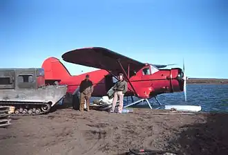 Two men standing facing camera under the wing of a red float plane. Some parcels of cargo are visible in the hatch, and a large tracked snow machine is parked on the left.