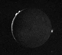 The thin crescent (open to the right) of the full disk of a planetary body with two bright clouds along the upper left edge of the object and another along the right edge.