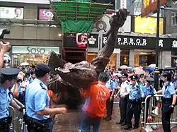 huge bronze sculpture being taken away by removal men dressed in orange, surrounded by police officers in blue shirts and caps or berets and overlooked by crowds