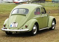 Picture of a yellow automobile in a field, with an airplane in the background. This is a rear three-quarters-view of it.
