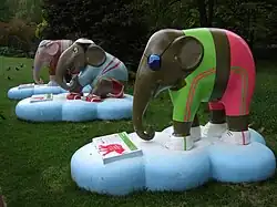 Three brightly coloured elephant statues line up on lush grass by trees in Hyde Park. All three elephants appear to be wearing sporting or athletic gear. Each statue stands on a light blue, cloud-shaped base, with an information plate displaying the artwork details.