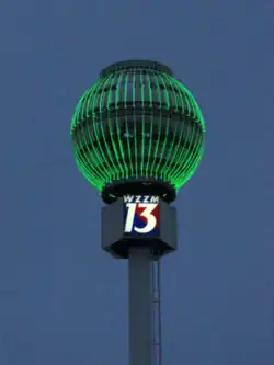 A large steel sphere with neon tubes lit in green, mounted high on a pole bearing the WZZM logo, at night