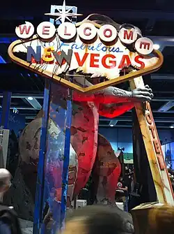 A damaged sign at says "Welcome to Fabulous New Vegas. An obscured dinosaur statue is behind the sign.