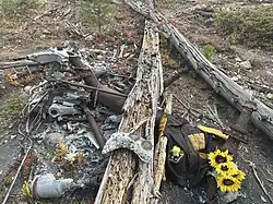 A large, rusted piece of aircraft machinery lies in a pile of twisted wreckage. A sticker on the machinery reads “Love Kansas.” A small religious statue is placed nearby, along with a yellow cloth draped over some of the wreckage. The area is surrounded by metal debris.