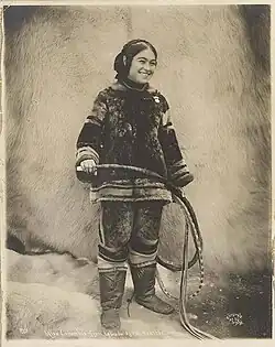 A smiling young Inuk woman, wearing traditional clothing, holding a whip