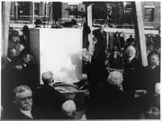 A black and white photograph of Secretary of State Charles Evans Hughes toweling mortar for the cornerstone of National Baptist Memorial Church. The stone is imposingly white in the center of the image and ringed by people wearing largely black, many of whom are turning towards the photographer.