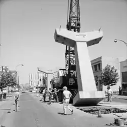 Construction crews look on as a mobile crane hoists a Y-shaped concrete column into the middle of a city street.
