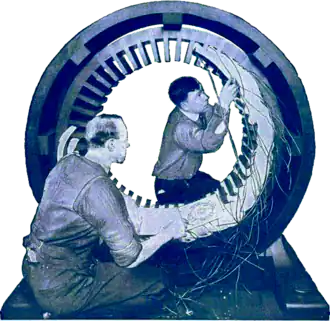 Blue-tinted photograph of two men working on a large circular piece of electrical equipment.