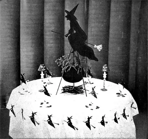 Black and white photograph of a small table decorated in a witch theme, the a central piece of a silhouetted witch on a broomstick over a cauldron.