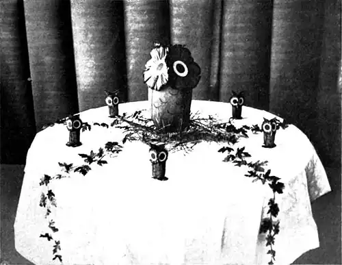 Black and white photograph of a small table decorated in an owl theme, the a central piece of a large owl, surrounded by little owls.