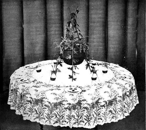Black and white photograph of a small table decorated in a witch theme, the a central piece of cauldron supported by three twigs and filled with foliage.