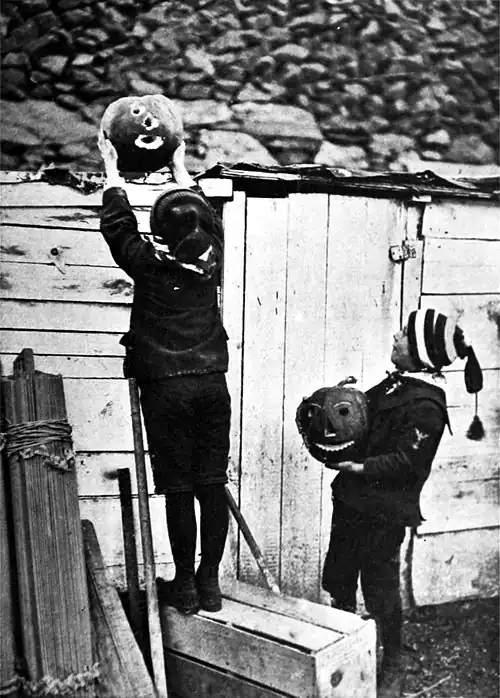 Black and white photograph of two boys with jack-'o-lanterns carved out of pumpkins. Oneis standing on a wooden box to place the lantern on top of a wooden shed.