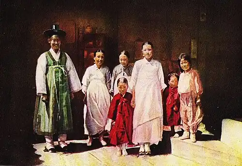 In this picture a Korean family is standing together. Three women are wearing dresses. Two boys are wearing long coats. One girl is wearing her pajamas. A fat man is wearing a bath robe, a rope around his chest, and a black cowboy hat that is too small for him.