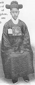 This picture is of a boy in a robe, white scarf, running shoes, and t-shaped hat. He is sitting with a tote bag on his lap. The tote bag is the same color as his robe. There is a folder sticking out of the bag with the picture of a lion or a poodle on it.