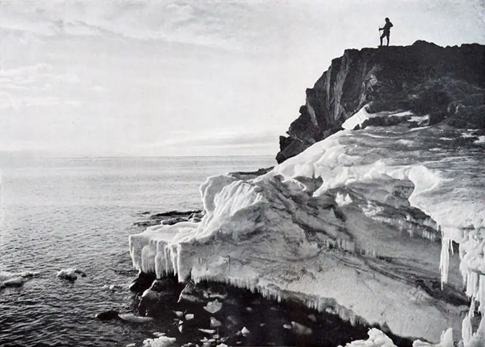Photo of a man standing atop a rocky cliff with ice at its foot, overlooking open water