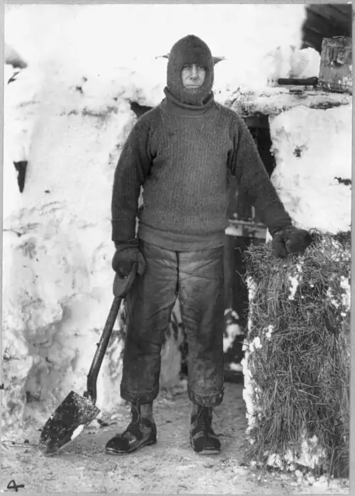 Full-length portrait of a man in a balaclava with one hand on a shovel and the other on a bale of hay
