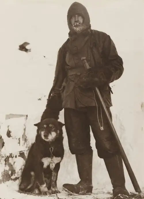 Full-length portrait of a man in outdoor gear with an alpenstock and a seated sled dog