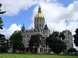 A color photograph of neo-gothic building with a golden dome on top of a central tower; trees, grass and a car park fill the foreground.