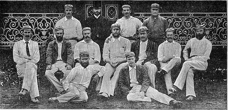 13 men dressed in cricket clothing posing for a group photograph. Three rows.