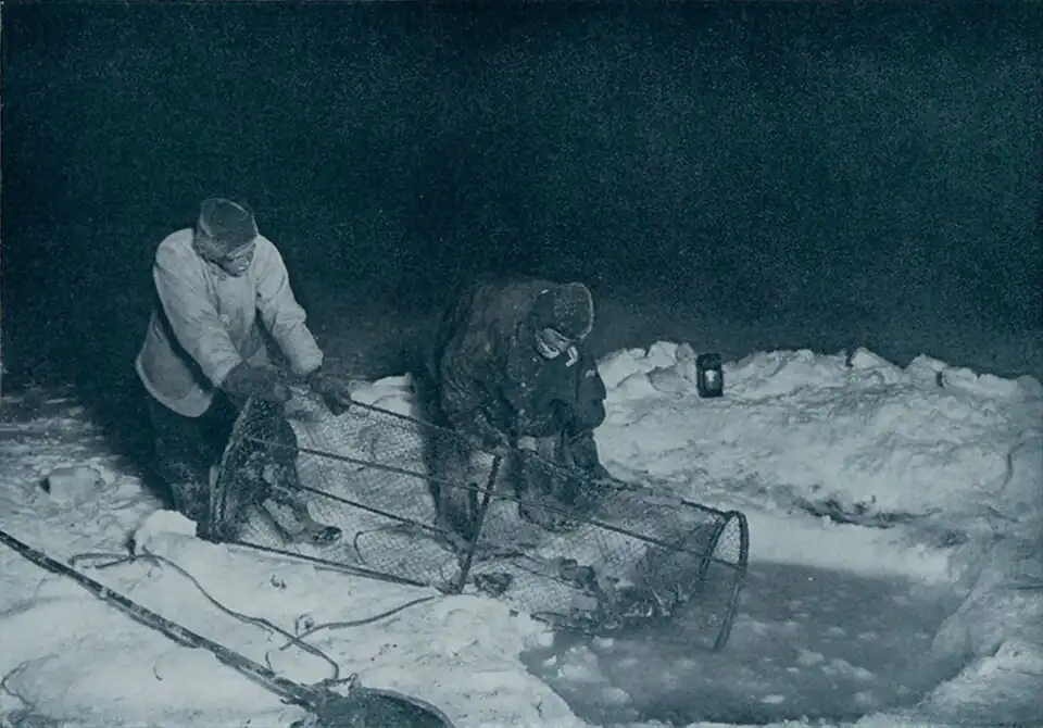 Photo of two men pulling a human-sized cylindrical cage out of a slushy hole in the ice