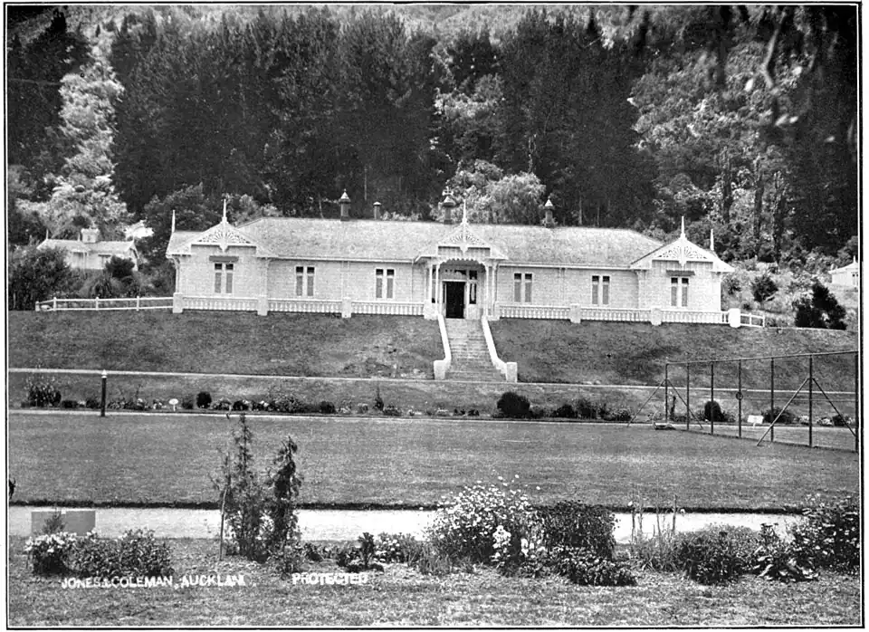 "Stately house with finials above window bays on the left and right hand side and the main entrance in the middle with steps in front; set on a terrace with trees in the background and lawns and garden on the lower terrace"
