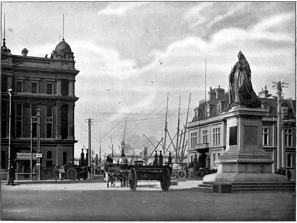 Statue of Queen Victoria on the right front, a horse and cart in the middle heading towards the wharf with ships masts visible, and two buildings on either side (the building on the left is Wellington Harbour Board Shed 7 (now the Queen's Wharf Apartments) and on the right is the 1982 Bond Store (now the Wellington Museum)