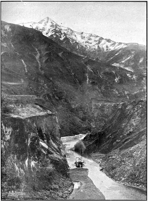A dredge in a a river surrounded by steep cliffs, barren hills, and a snow-topped mountain