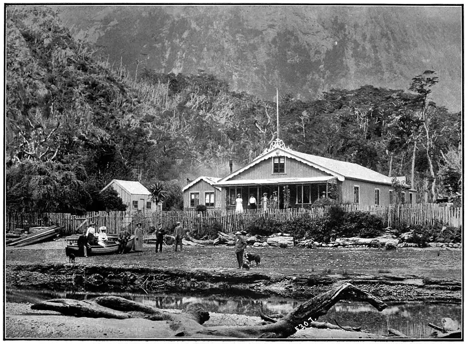 Large house with a covered verandah at the front and additional buildings, bush covered hills in the background and a shore in front with a small boat